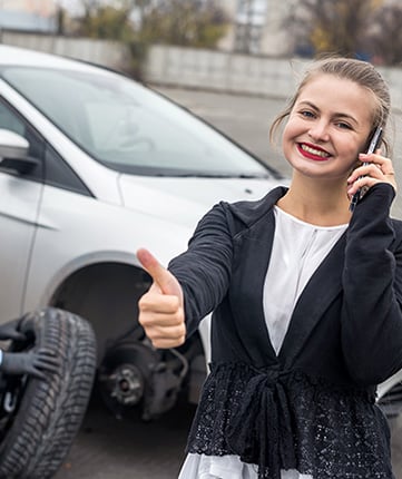 Woman standing by broken down with flat tire, on her mobile phone with a thumbs up